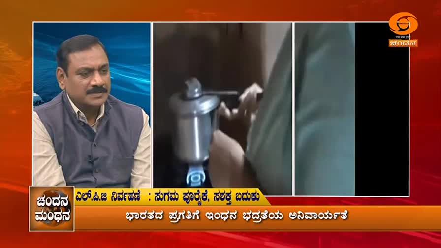 A man with a mustache sits in a studio, looking thoughtfully to his left. To his right, a hand turns a valve on a metal appliance, a blue flame visible beneath it.