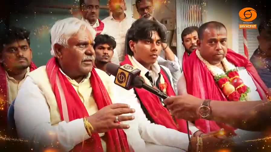 An older man, his white hair contrasting with a bright red shawl, speaks into a microphone held by an unseen interviewer. Beside him, a younger man in a white shirt also wears a red shawl, his gaze fixed forward.