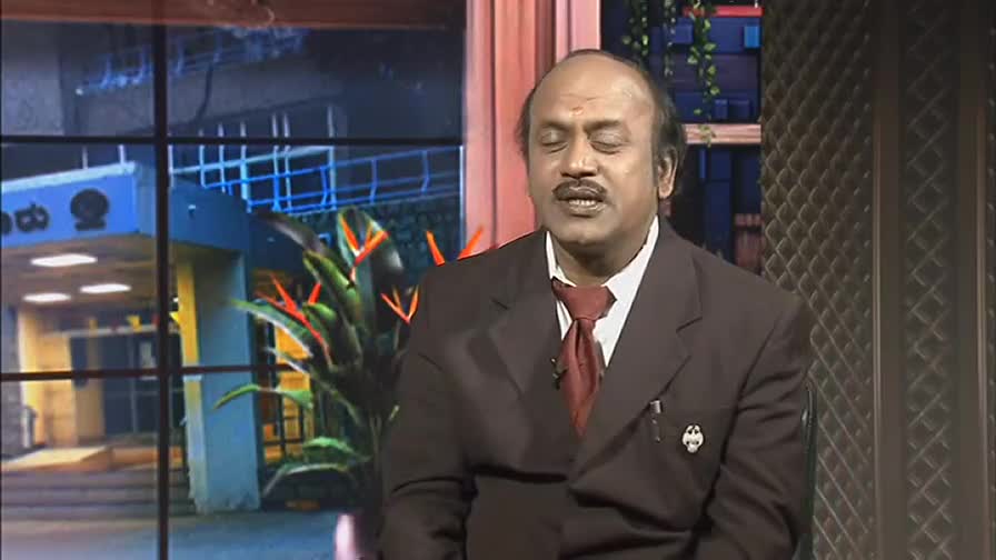 A man in a suit speaks with his eyes closed, his expression earnest. Behind him, a studio backdrop displays a building with Kannada script and bright orange flowers.