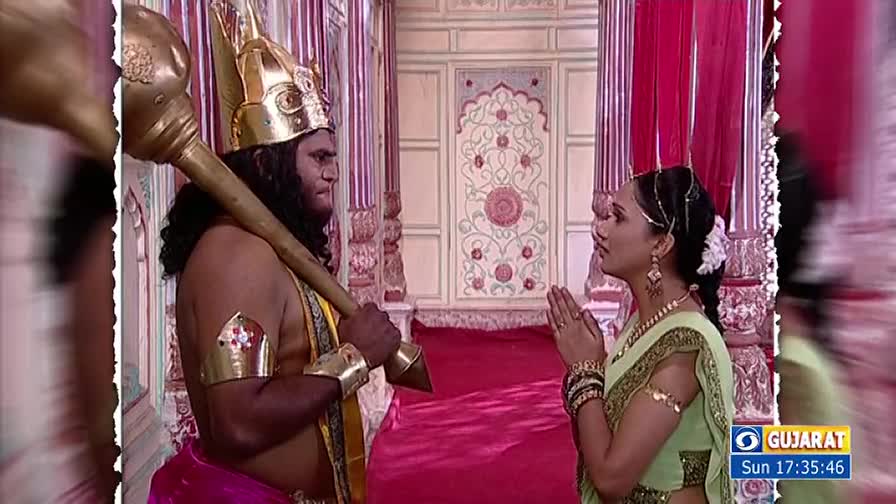 A man in a golden helmet and armbands stands with a large club, facing a woman with her hands pressed together. She is dressed in a sari and looks at him with a respectful expression.
