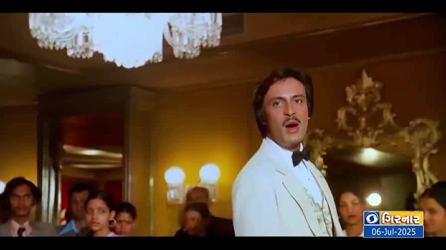 A man in a white suit and bow tie is singing, his mouth open in mid-note. Behind him, a crowd watches, and the DD Girnar logo is in the corner.
