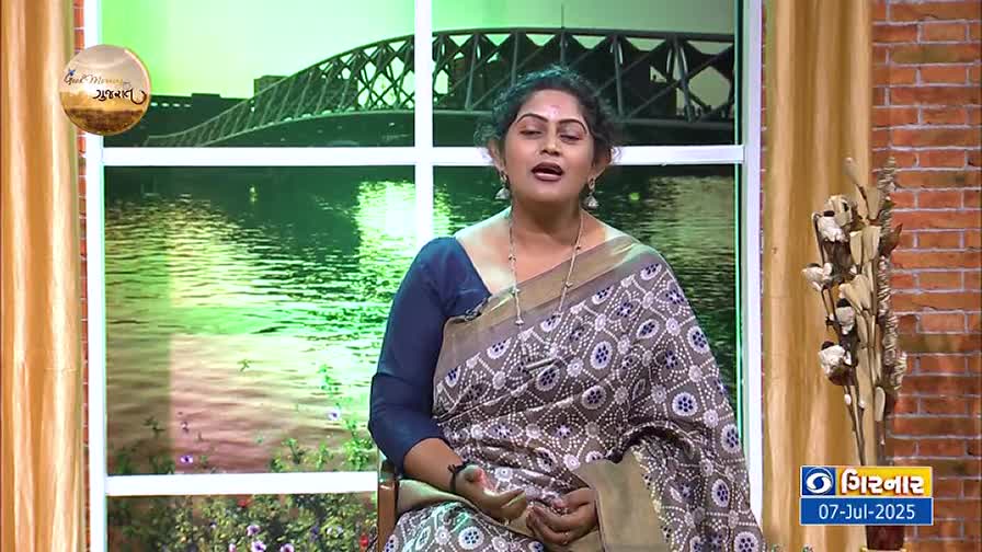 A woman in a patterned sari sits in a studio, speaking directly to the camera. Behind her, a backdrop shows a bridge over water, and the DD Girnar logo is visible on the lower right.
