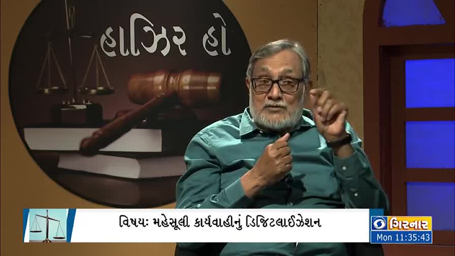 A man with a gray beard and glasses gestures with his right hand while speaking. Behind him, a logo for DD Girnar is visible, along with a gavel and scales of justice.
