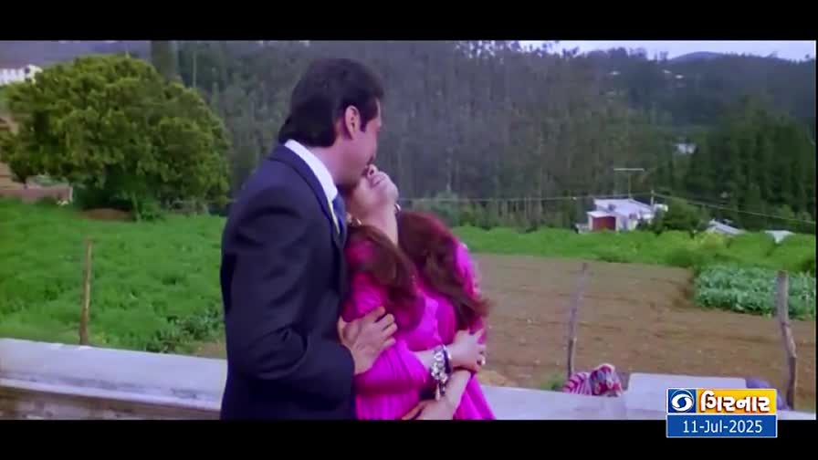 A man in a dark suit embraces a woman in a bright pink dress, their faces close. The scene is set against a backdrop of green hills and a DD Girnar logo in the corner.
