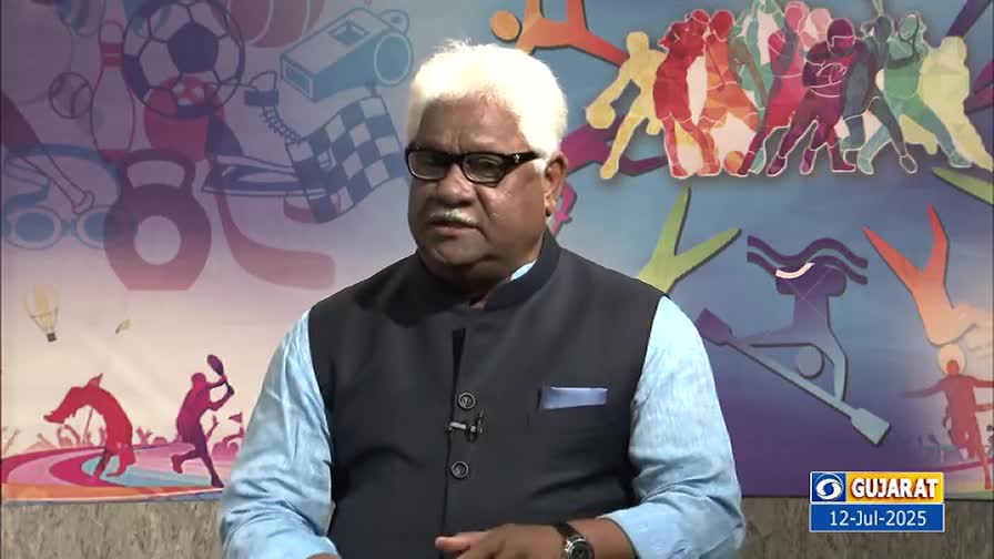 A man in a dark vest and glasses is speaking, his gaze directed off-camera. Behind him, a colorful backdrop features silhouettes of various sports figures.
