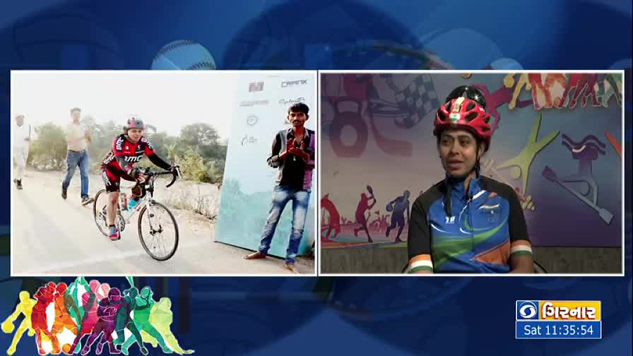 A cyclist in a red and black outfit pedals past a sign, while a man watches. On the right, a woman in a helmet sits, speaking on the DD Girnar broadcast.

