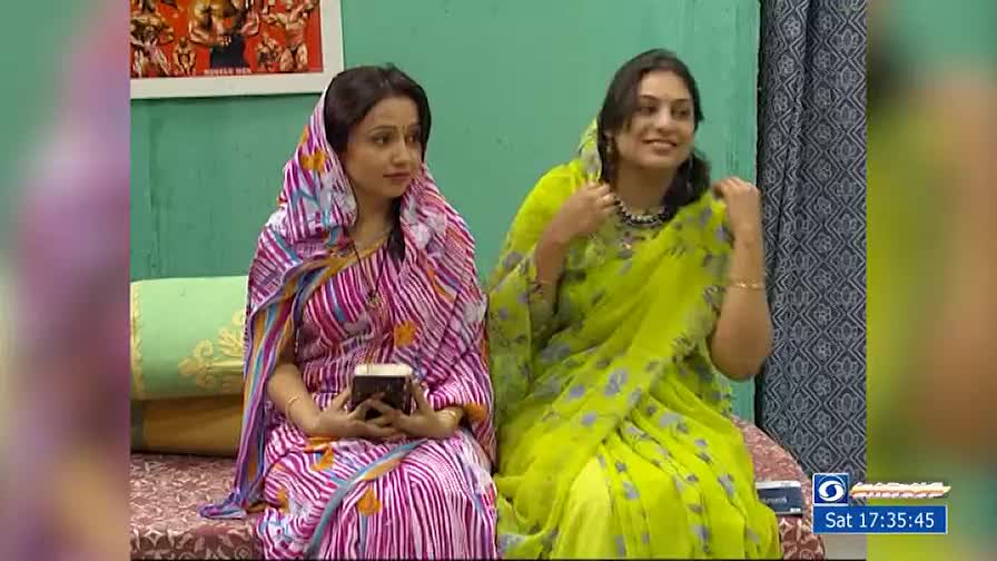 Two women in bright saris sit side-by-side, the one on the left holding a small, patterned box. The woman on the right adjusts her necklace, smiling at someone off-camera.
