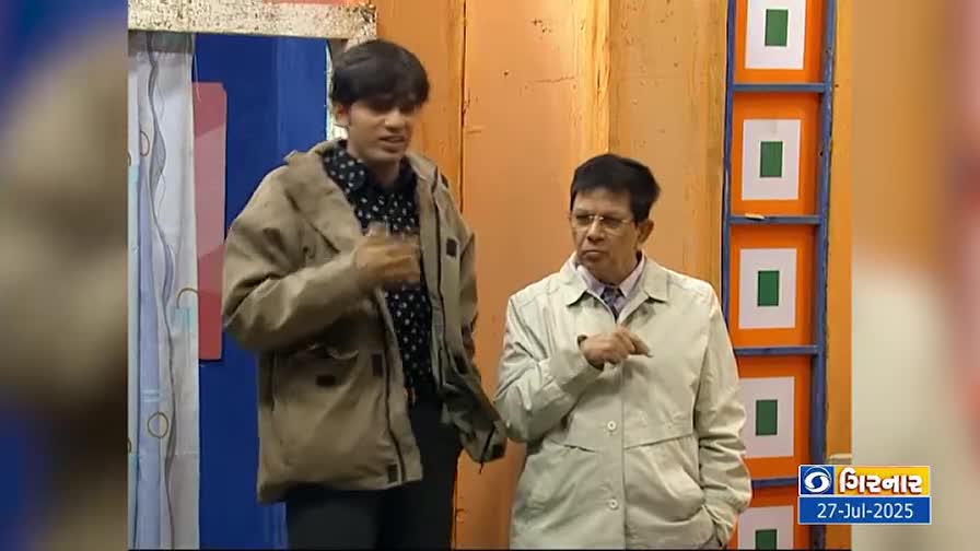 A younger man in a tan jacket gestures emphatically, speaking to an older man who listens intently. The backdrop is a simple set with painted panels and a DD Girnar logo in the corner.
