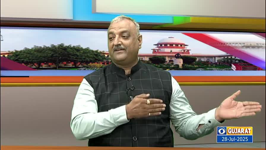 A man in a vest and light-colored shirt gestures with his hands, speaking in front of a backdrop of a building. The screen behind him shows a building that looks like the Supreme Court of India.
