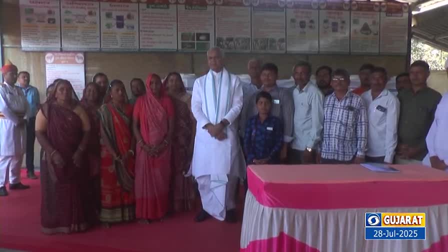A group of people, many in traditional Indian dress, stand facing the camera. A man in white, with a white scarf, is in the center, and a table draped in pink sits in front of them.
