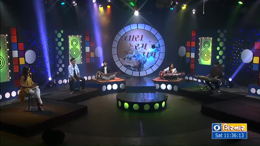 On a brightly lit stage, musicians with stringed instruments are seated, ready to perform. A woman in a yellow top sits to the left, while the DD Girnar logo is displayed in the bottom right corner.
