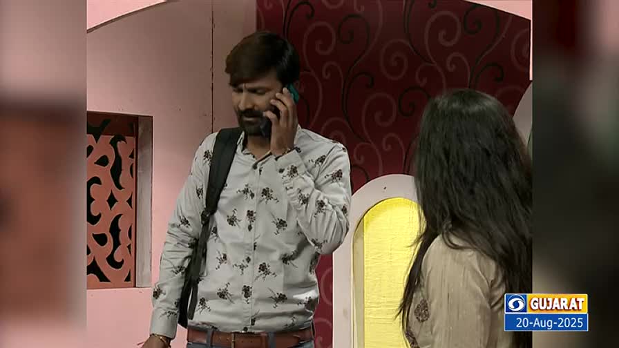 A man, wearing a floral shirt and a backpack, is talking on a phone, his gaze directed downward. Beside him, a woman stands with her back to the camera, the scene framed by a decorative wall and the DD Girnar logo.

