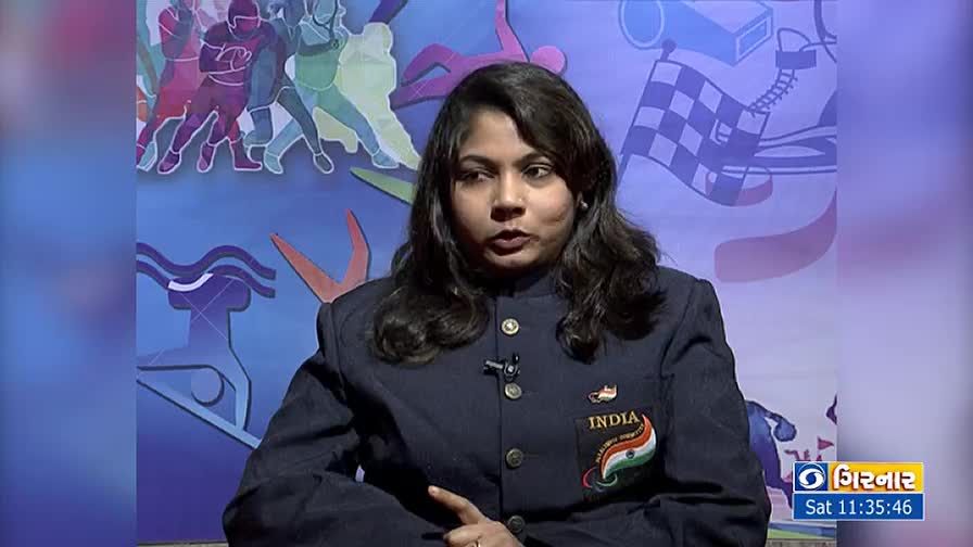 A woman in a navy blazer with an "INDIA" patch is speaking on camera, her hand gesturing. Behind her, a backdrop with sports-themed illustrations and a logo for DD Girnar is visible.
