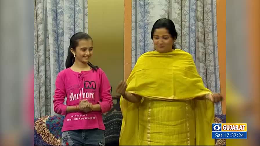 A woman in a bright yellow outfit gestures with her hands, while a girl in a pink shirt smiles beside her. The scene is set in what looks like a television studio, with the DD Girnar logo visible.
