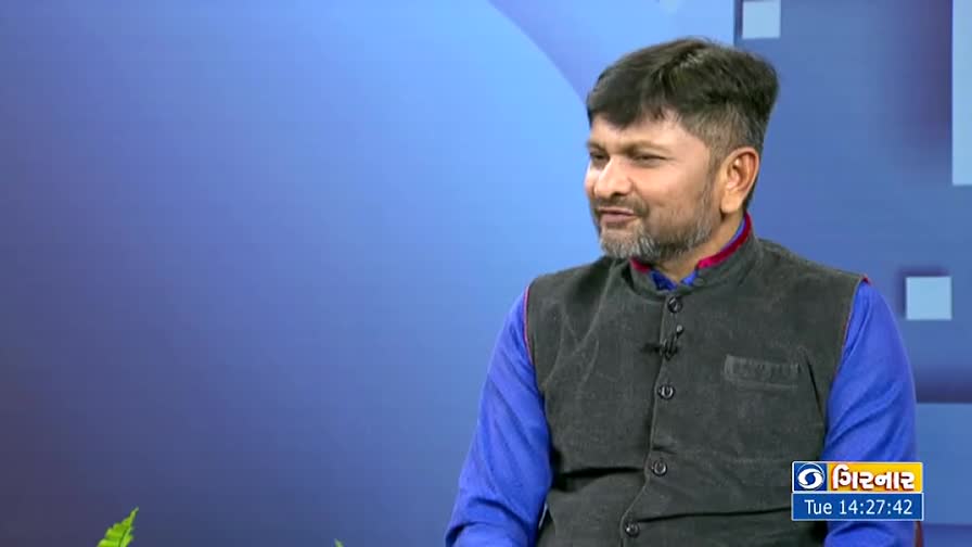 A man with a trimmed beard and dark vest sits in front of a blue backdrop, speaking. The DD Girnar logo is visible in the corner of the screen.
