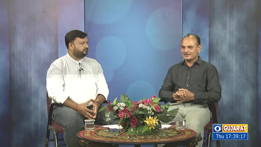 Two men sit across from each other at a small table, a bouquet of flowers between them. One wears a white shirt, the other a dark one, both looking forward, as if in conversation.
