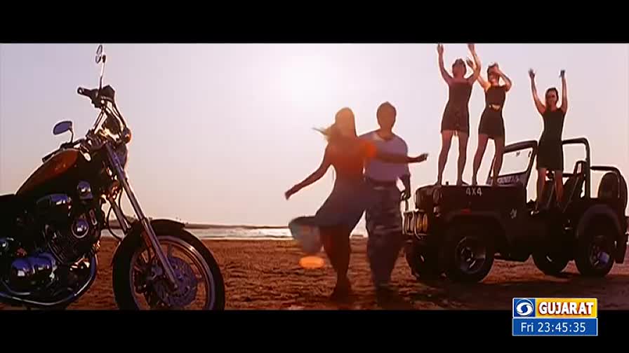 A shiny motorcycle sits parked on the sand as a couple dances nearby. Three women stand on top of a jeep, arms raised in the air.
