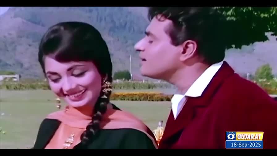 A woman in a braided hairstyle smiles, and a man in a maroon jacket turns his head toward her. They're outside, with green grass and mountains in the background.
