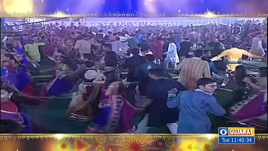Dancers in bright clothing move in a large circle, some with drums, as the crowd sways to the music. The scene is lit with bright lights, and the energy is palpable.
