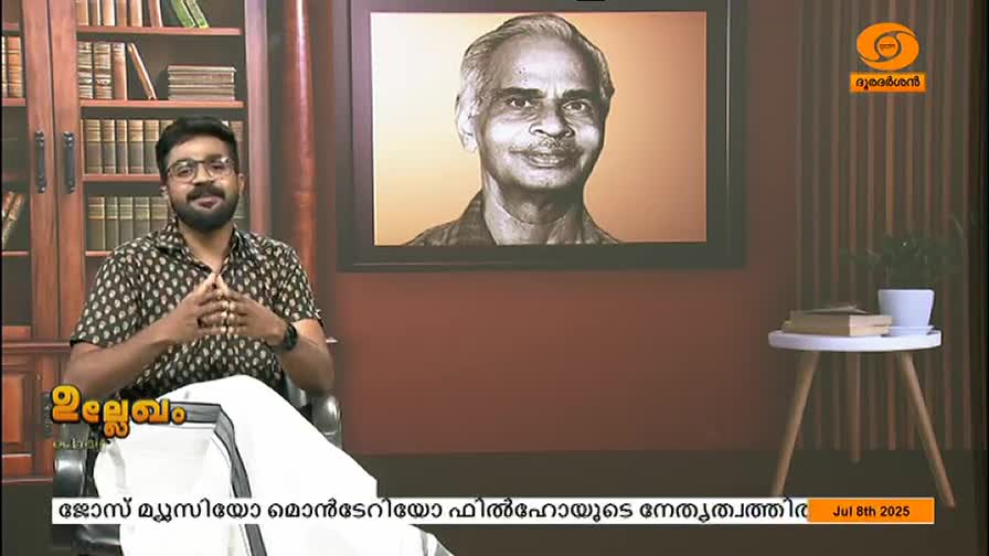 A man in glasses and a patterned shirt sits in a studio, gesturing with his hands. Behind him, a portrait hangs on the wall, and the DD Malayalam logo is visible in the corner.
