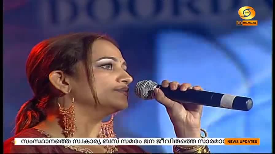 A woman, lit by stage lights, holds a microphone close to her mouth. The DD Malayalam logo is visible in the corner.
