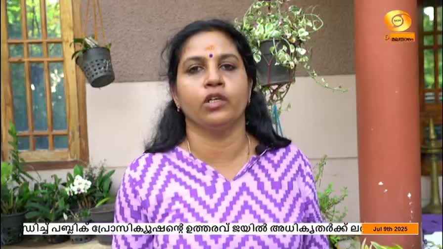 A woman in a purple patterned top speaks directly to the camera, a small microphone clipped to her collar. Behind her, plants hang in pots, and a logo for DD Malayalam is visible.
