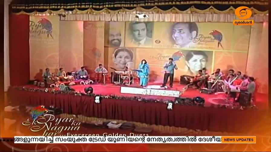 A woman in a blue dress sings, while a man in a blue shirt gestures to the audience on a stage decorated with portraits. Musicians play instruments behind them, as the DD Malayalam logo is visible.
