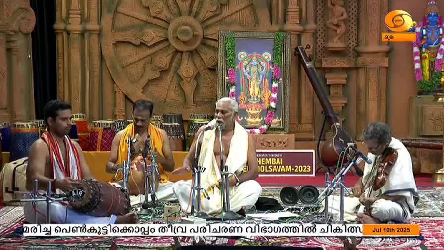 A singer, dressed in white, sits center stage, holding a microphone and gesturing with his hands. Musicians surround him, playing a variety of instruments, including a violin and percussion, as part of the DD Malayalam broadcast.
