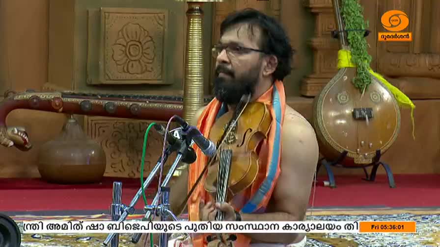 A man with a beard and glasses holds a violin, bow poised, ready to play. He's on a stage, with a traditional Indian instrument behind him, and a DD Malayalam logo in the corner.
