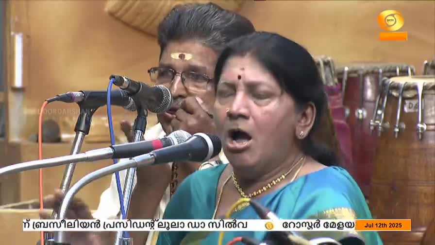 A woman in a teal sari sings into a microphone, eyes closed, her mouth wide open. Beside her, a man with glasses and a white shirt leans in, also singing, as the DD Malayalam broadcast continues.

