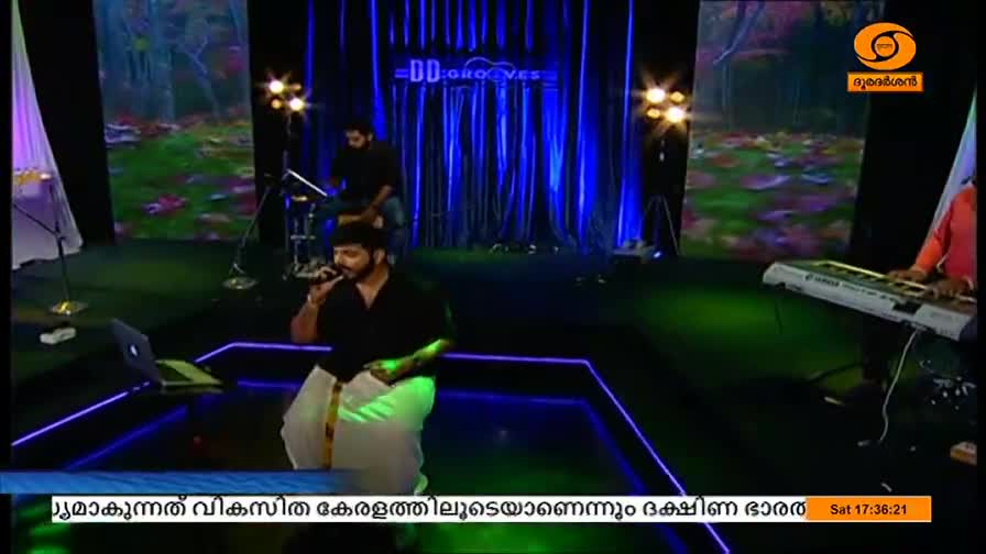 A man in a black shirt and traditional white mundu sings into a microphone, a laptop open in front of him. Behind him, a drummer and keyboard player provide the music on a stage lit with blue and green lights, likely for DD Malayalam.
