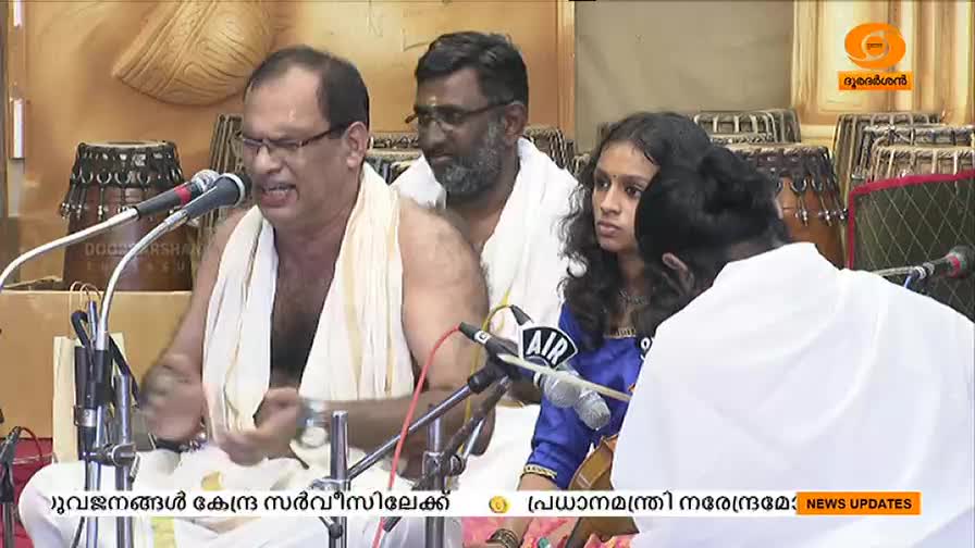 A man in a white cloth speaks into a microphone, his expression animated. Seated beside him, a young woman listens intently, while other men look on.
