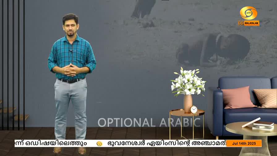 A man in a blue plaid shirt stands in front of a gray backdrop, hands clasped. Behind him, a blurred image suggests a scene from India, possibly a news report on DD Malayalam.
