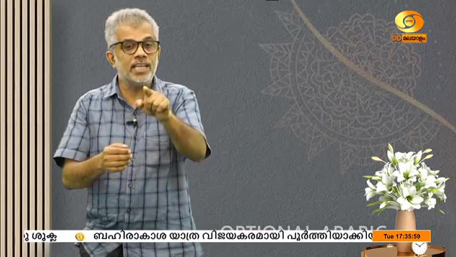 A man in glasses gestures with his right index finger, speaking animatedly on a DD Malayalam set. The background is a muted gray, with a floral arrangement and a clock visible to the right.
