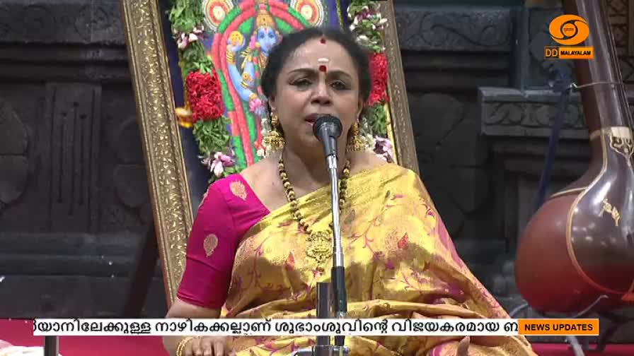 A woman in a vibrant sari sings into a microphone, her voice filling the space. Behind her, a framed image of a deity and a stringed instrument stand as silent witnesses.
