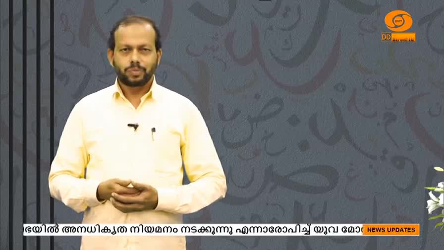 A man in a light yellow shirt stands centered, looking directly at the camera. The DD Malayalam logo is visible in the upper right corner, and text scrolls along the bottom of the screen.
