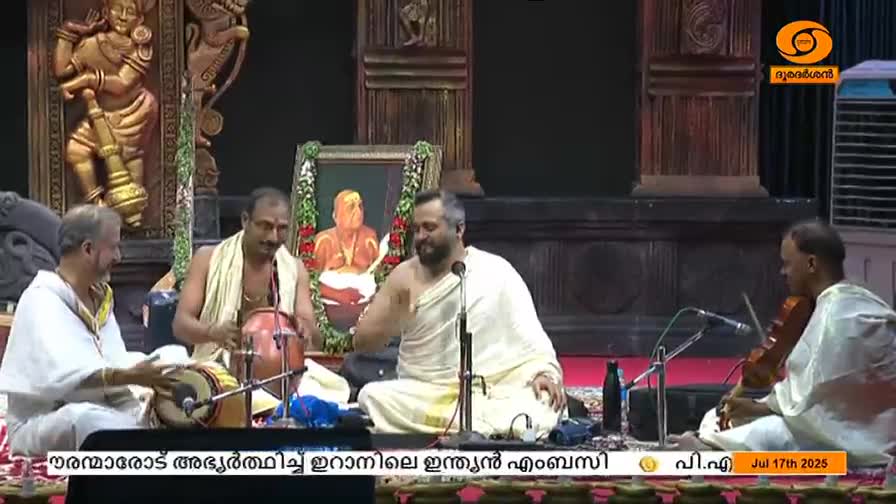 A man in white robes gestures with his right hand as he sings into a microphone. The musicians around him, including someone playing a violin, are focused on the music.

