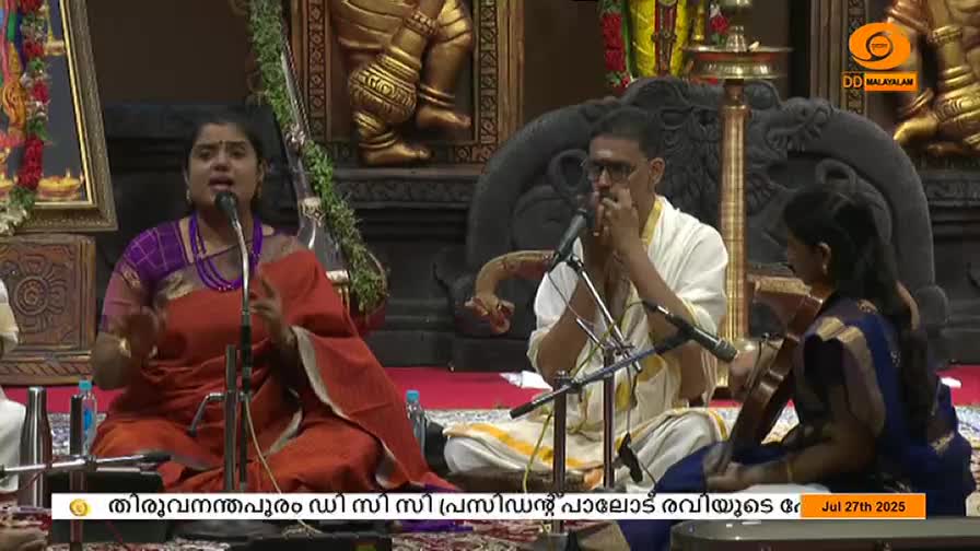 A woman in a red sari sings into a microphone, gesturing with her hands. A man plays a wind instrument, while a young woman accompanies on the violin, all seated on the floor in front of a DD Malayalam backdrop.

