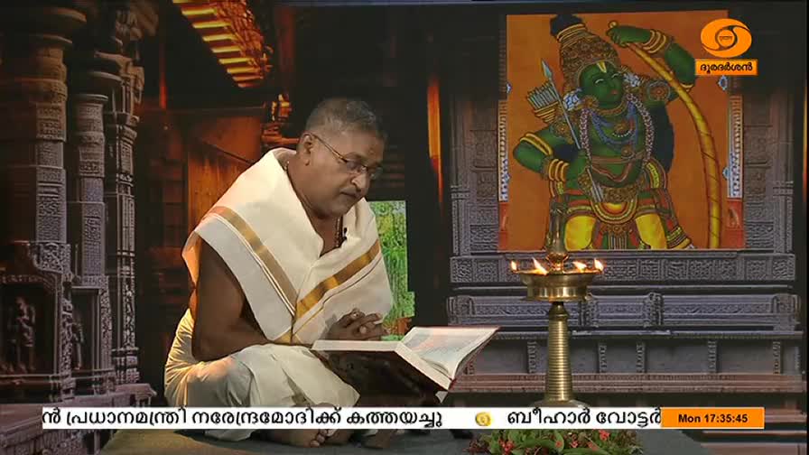 A man in traditional white attire sits cross-legged, reading from an open book. Behind him, a painting of a green-skinned figure with a bow and arrow hangs on the wall, a traditional oil lamp glowing in front of it.
