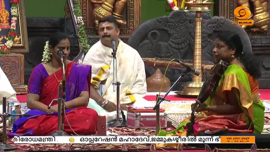 A woman in a green and orange sari plays a stringed instrument, drawing a bow across the strings. Two other performers sit nearby, one singing into a microphone, the other looking on, all on a stage with the DD Malayalam logo visible.
