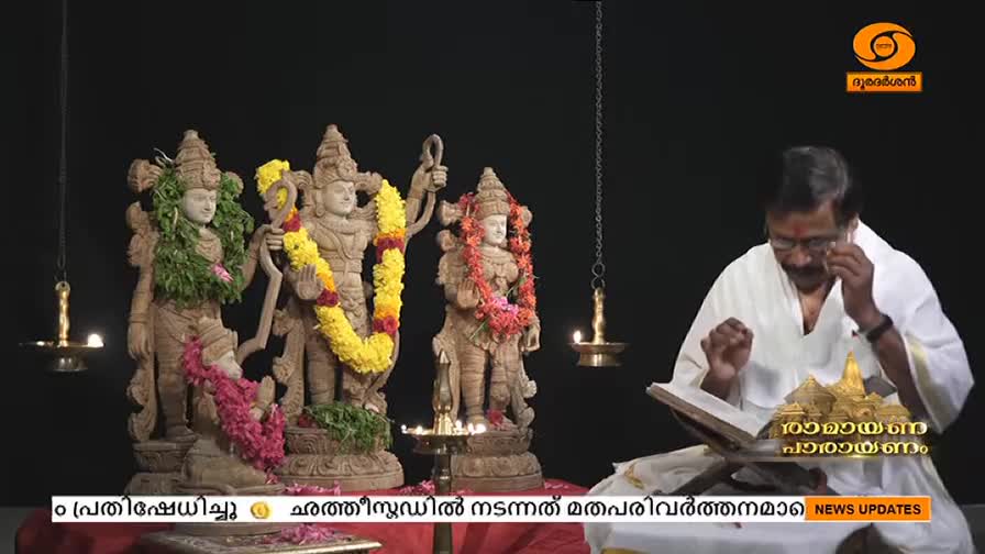 A man in white robes reads from a book, lit by a hanging lamp, with statues of deities adorned with garlands behind him. The DD Malayalam logo is visible, and the scene is likely part of a religious broadcast from India.
