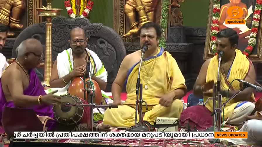 A group of musicians, dressed in traditional attire, are performing on a stage. The man in the center, wearing a yellow garment, sings into a microphone as the others play various instruments.
