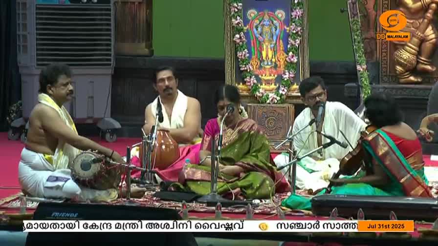 A man in a white dhoti and shawl strikes a drum, while others sit cross-legged, ready with their instruments. A woman in a green sari bows her head and appears to be singing into a microphone.
