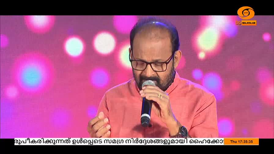 The man in the coral shirt holds a microphone, singing with his eyes closed. The DD Malayalam logo is in the upper right corner, and the background is a soft pink with blurred lights.
