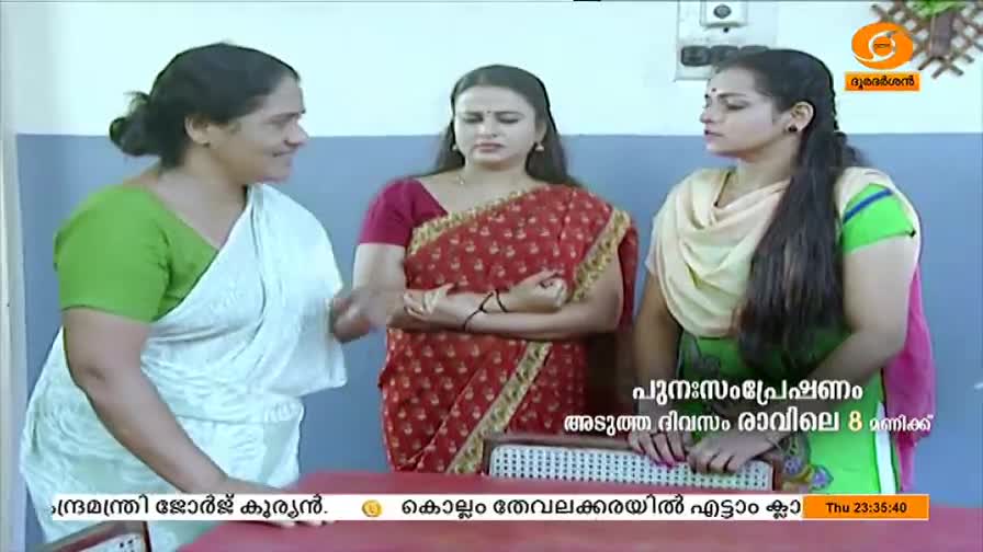 Three women stand together, the one on the left gesturing with her hand as she speaks. The woman in the middle, wearing a red sari, looks on with a serious expression.
