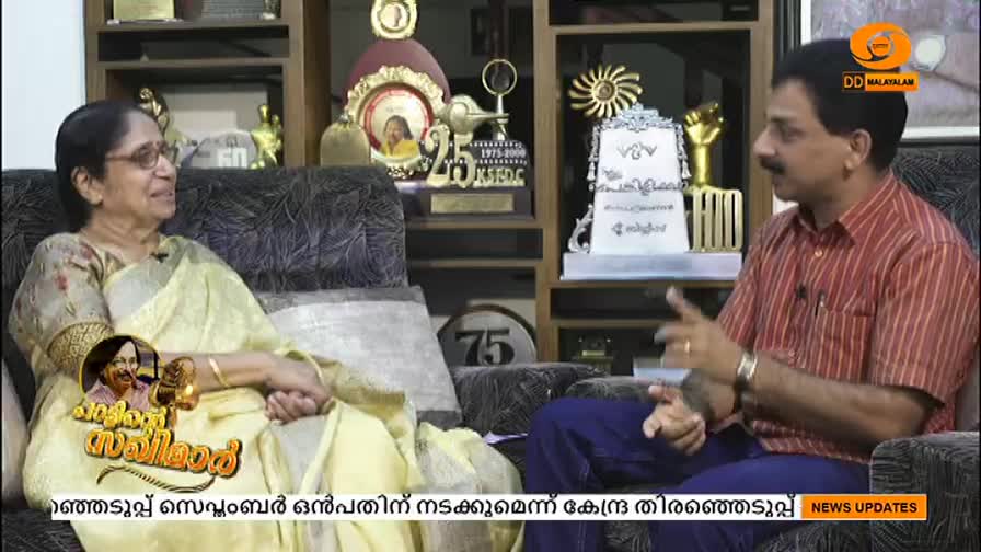 A woman in a yellow sari sits on a couch, smiling as a man in a red shirt gestures while speaking. The scene appears to be a DD Malayalam interview, with a backdrop of awards and trophies.
