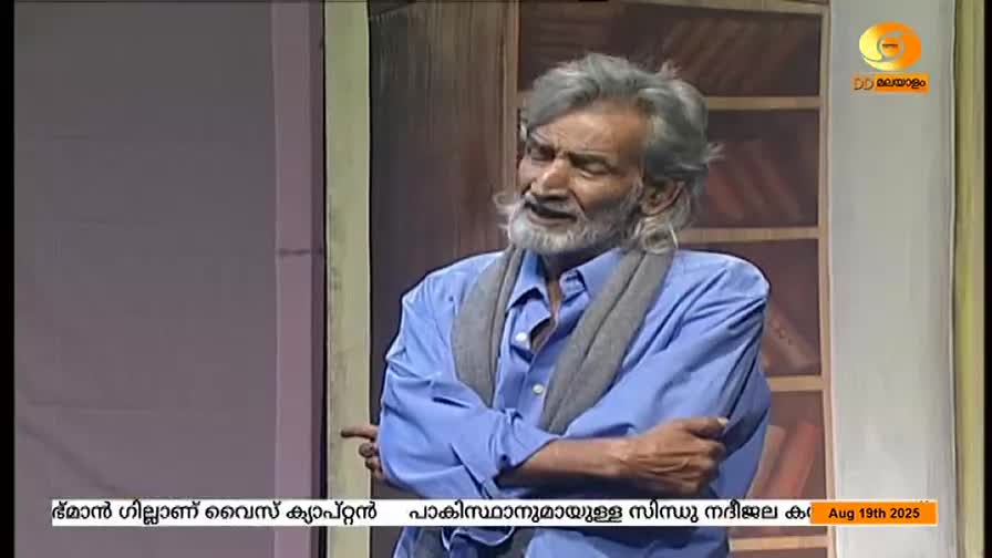 An older man in a blue shirt gestures while speaking, his face showing emotion. The DD Malayalam logo is visible in the corner, and text scrolls at the bottom of the screen.

