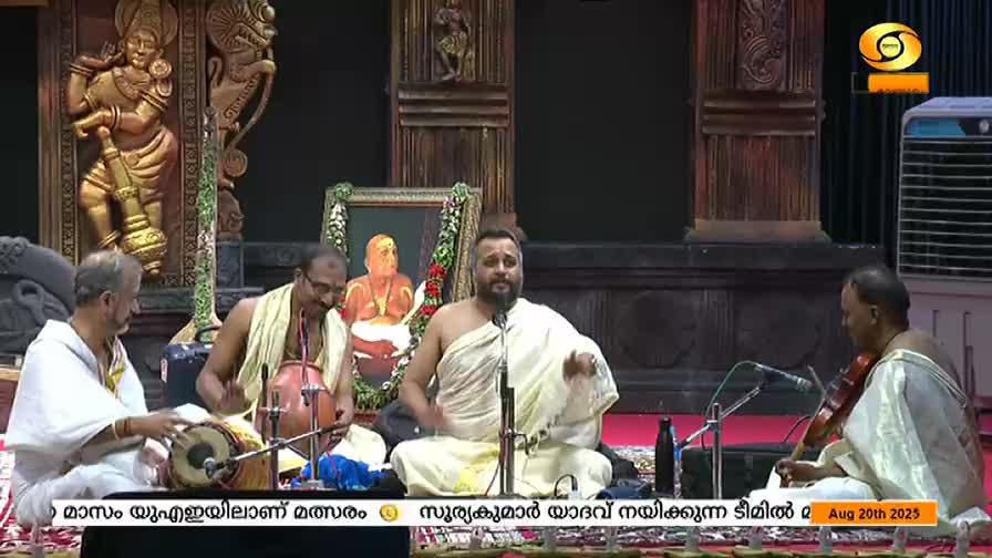 A vocalist in white robes gestures as he sings, while musicians accompany him on a mridangam, a violin, and a stringed instrument. The DD Malayalam logo and text at the bottom indicate this is a broadcast from India.

