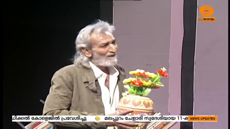 An older man with long, white hair and a beard holds a small, decorated pot with colorful flowers. He appears to be speaking on a DD Malayalam news program, with text scrolling at the bottom of the screen.
