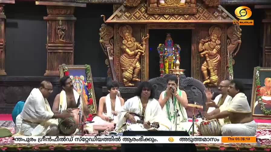 Musicians in white robes are seated on the floor, some playing instruments like drums and a stringed instrument, while others sing. The DD Malayalam logo is visible, and the backdrop is a decorated stage with religious figures.
